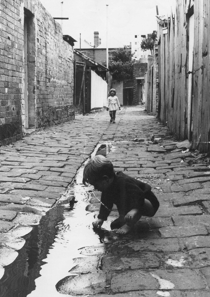 This is a black and white photo of a young boy playing in a puddle in a Fitzroy laneway.