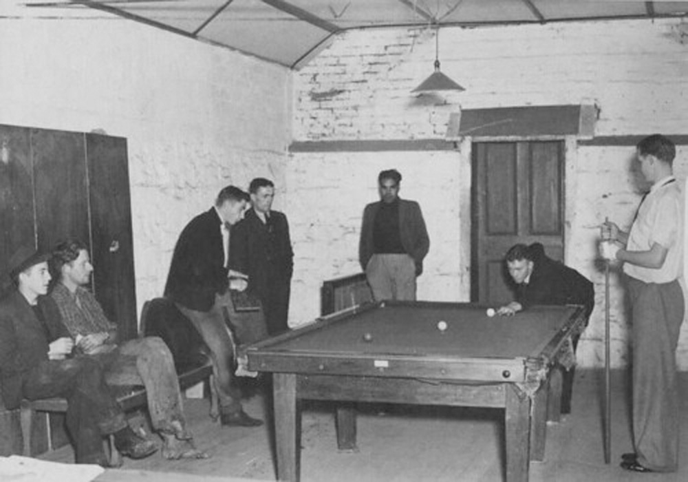 This black and white photograph is of seven young men playing billiards and sitting around the table in the club room at the boys hostel in Fitzroy.