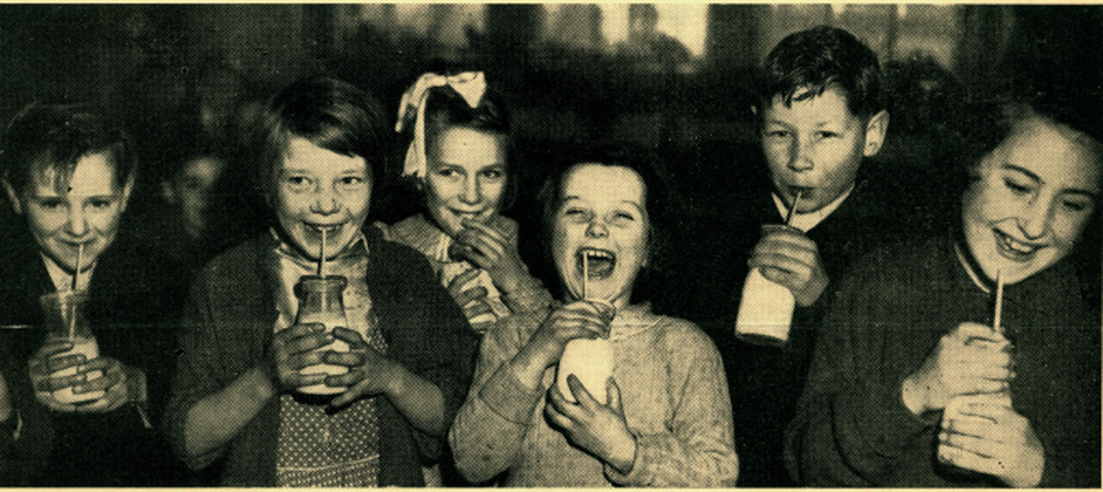 This is a black and white photograph of children enjoying free milk at school There are about 8 children in the photo and they are happy and smiling.
