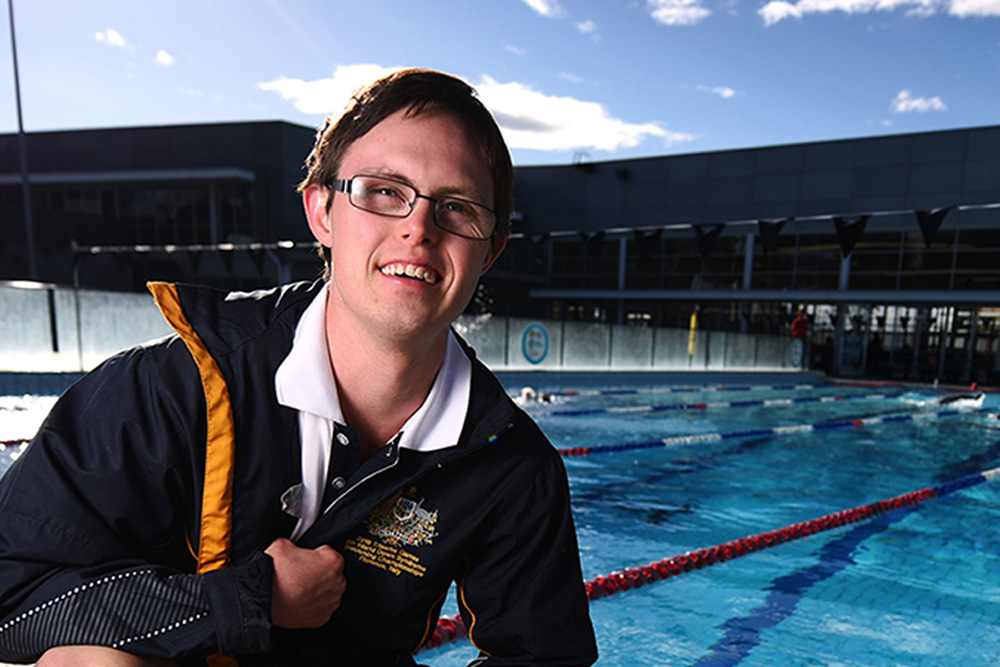 This is a photo of an NDIS Participant standing with his bag and smiling at the camera. He is in front of a pool.