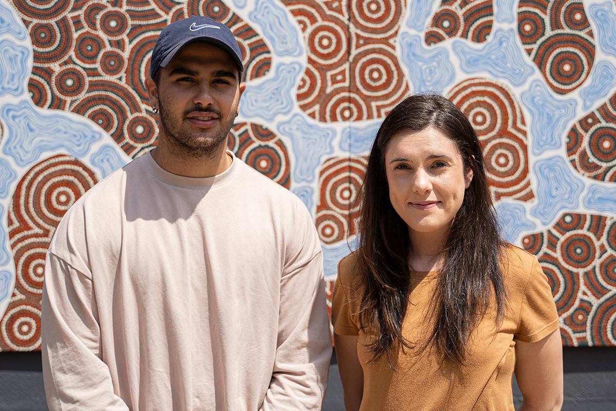 This is a photo of CareerTrackers interns Stephany and Jamarl undertook internships with our youth programs. They are smiling and the camera and are standing in front of Aboriginal artwork.