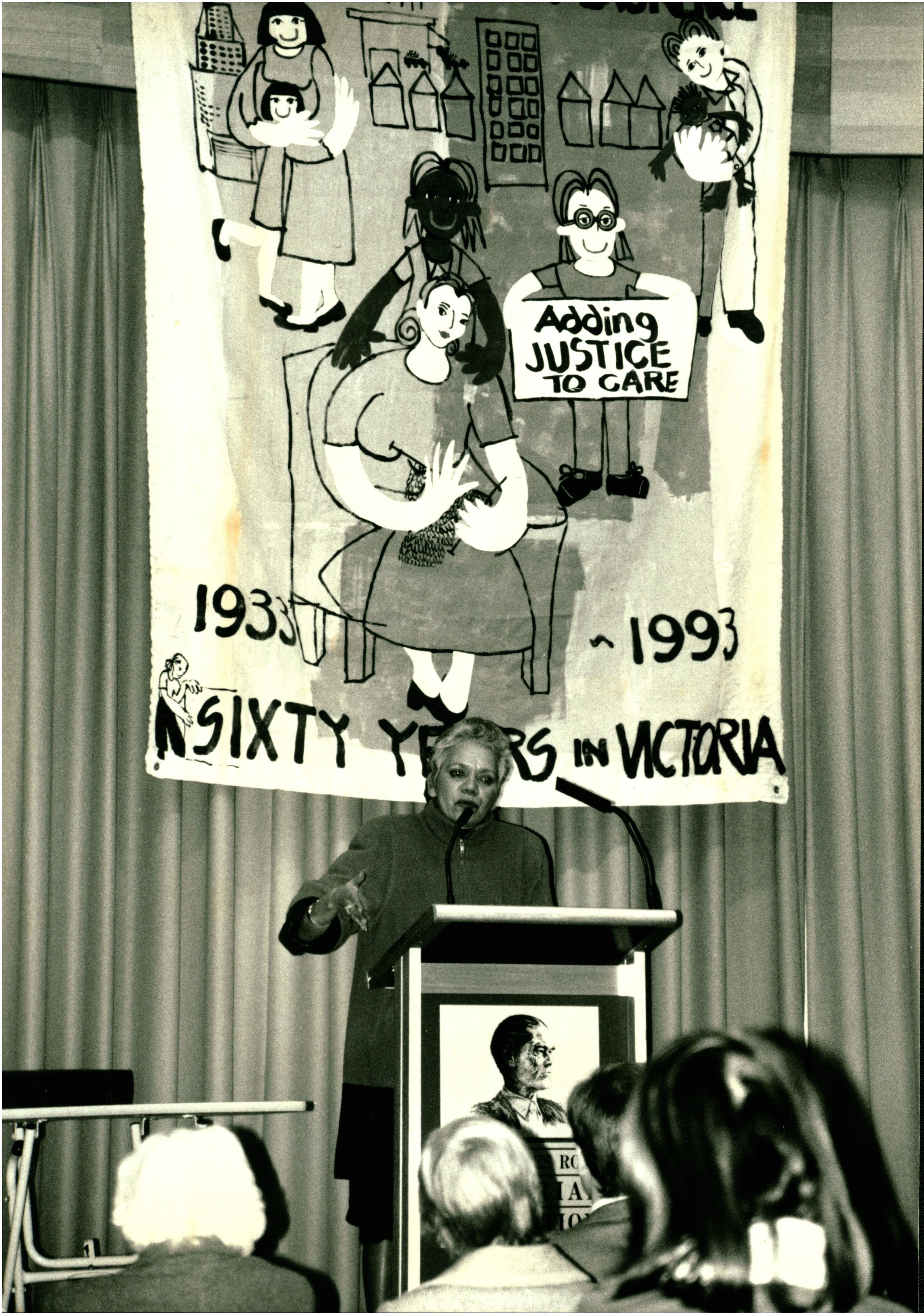 This is a black and white photo of Lillian Holt addressing the audience at the 1993 Sambell Oration. She is standing in front of a banner that says 'Adding justice to care' and '60 years in Victoria'