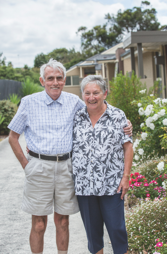 This is a photograph of an older couple standing outside the Banksia Centre. There are gardens in the background. The couple is smiling.